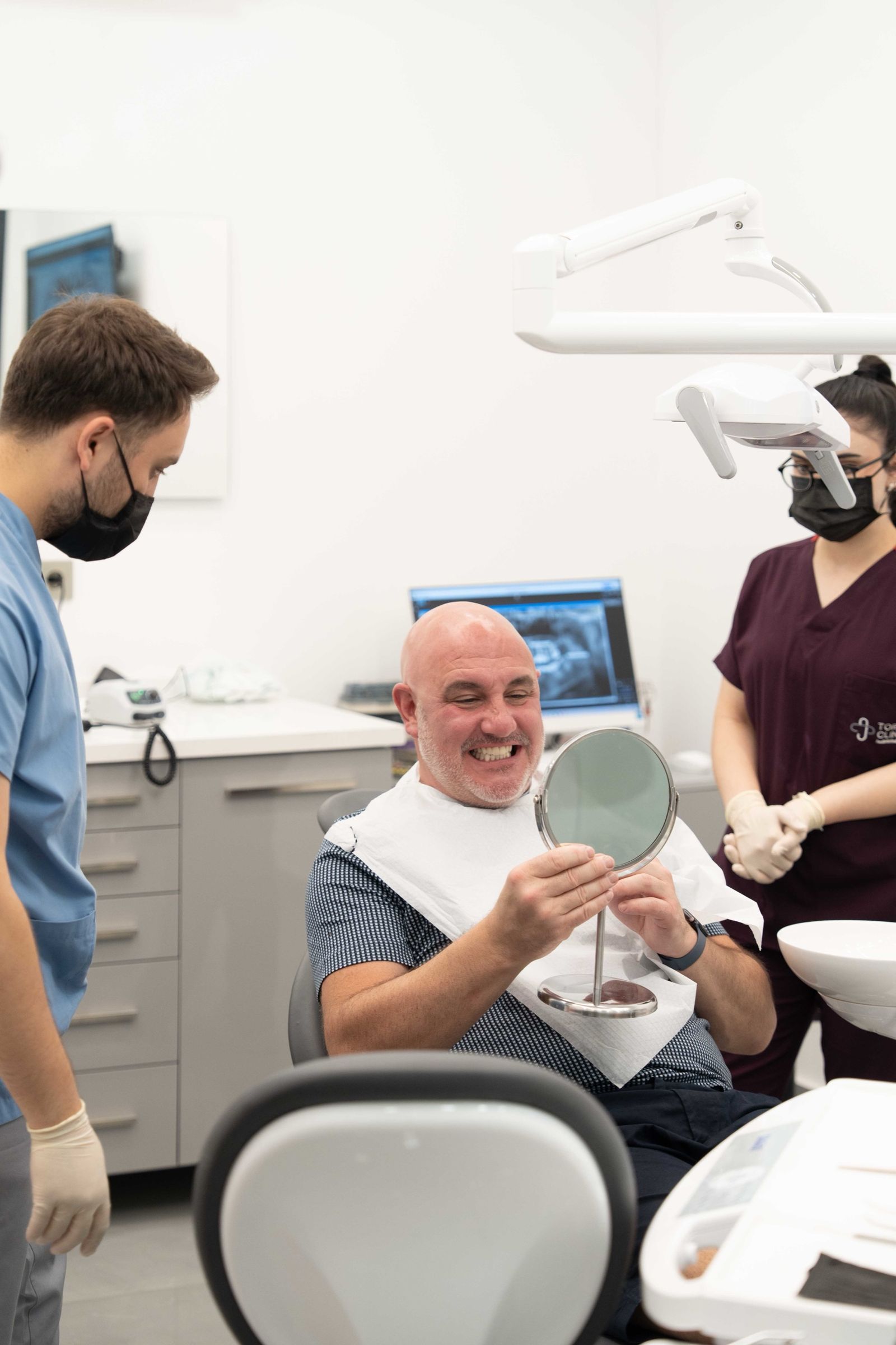 A patient smiling at his reflection in a hand mirror, with the dental team beside him.